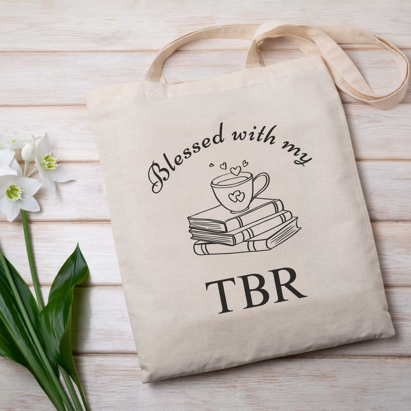 Natural tote bag with 'Blessed with my TBR' text & book graphics, placed on a wooden surface with flowers, bookish gift for readers.